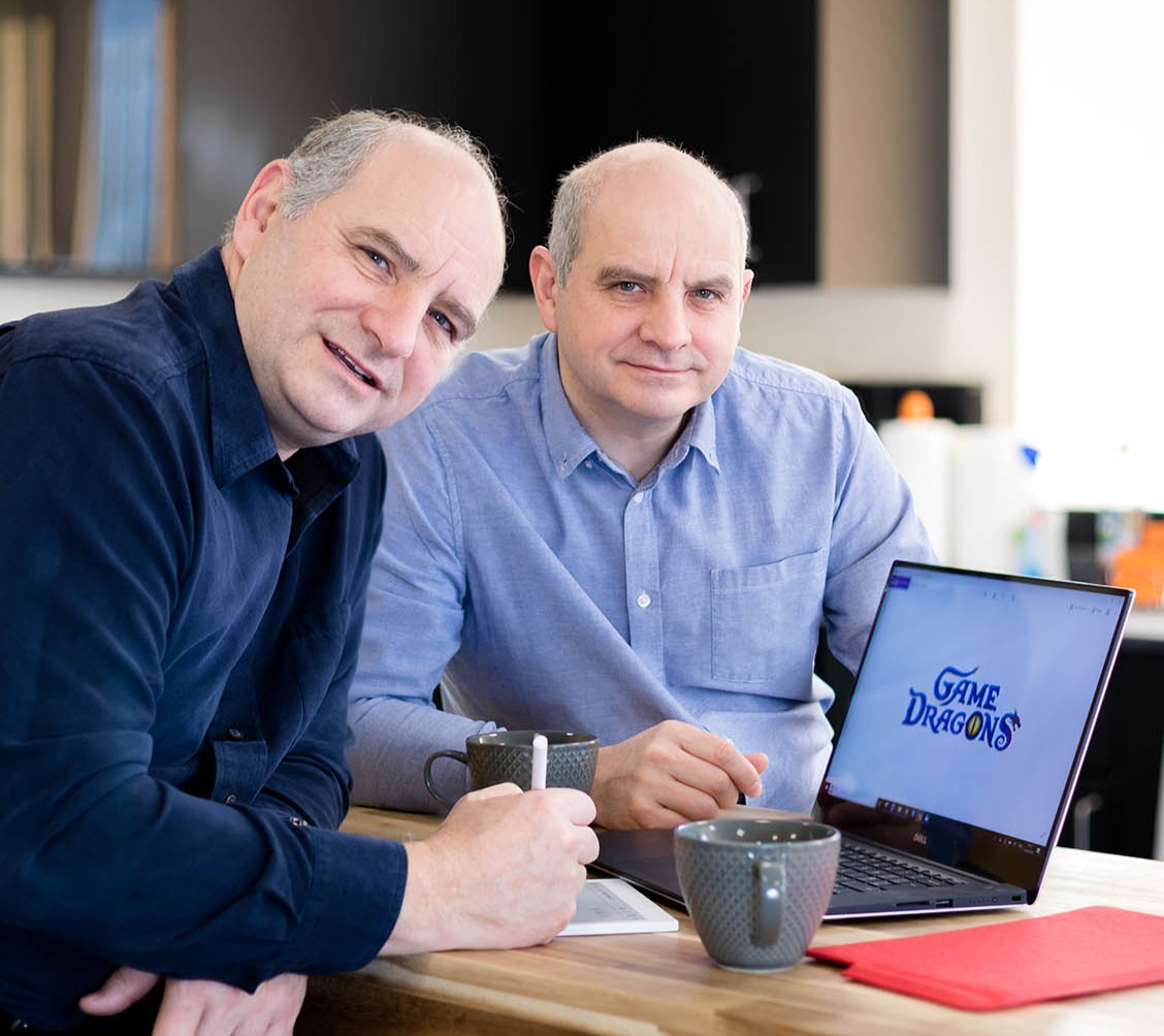 Philip und Andrew Oliver am Tisch mit einem Notebook mit Game-Dragons-Logo - Bild: Game Dragons